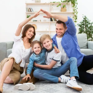 A husband and wife with their children, a boy and a girl, sitting on the floor smiling together. The parents are holding their arms up together, making the shape of a roof of a house